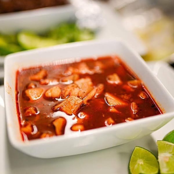 A white bowl of red stew with meat chunks, accompanied by lime wedges and green chilies.