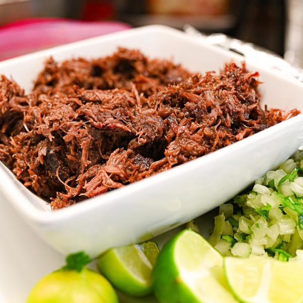 Shredded beef in a white bowl, with chopped onions, cilantro, and lime wedges on the side.