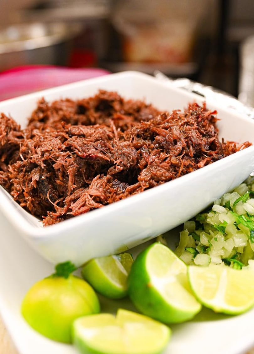 Shredded beef in a white bowl, with chopped onions, cilantro, and lime wedges on the side.