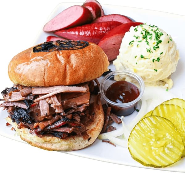 A BBQ brisket sandwich with sides of sausage, mashed potatoes, pickles, and onions.